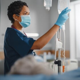 Hospital room with young dark-complected nurse wearing face mask checking on patient's IV fluid; patient is blurred with gray hair.